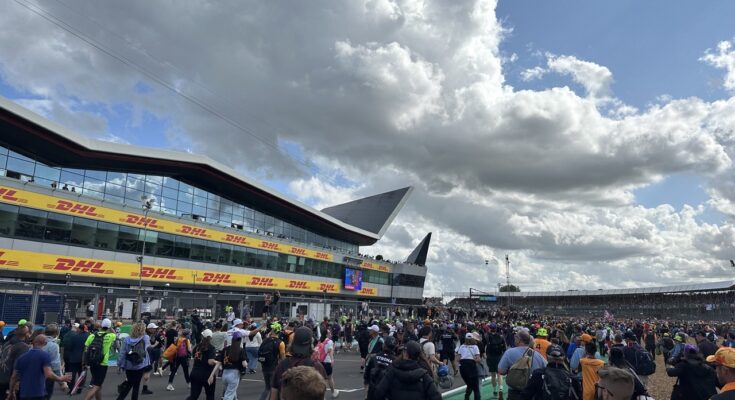 Fans running down main straight at Silverstone towards podium, with a blue cloudy sky above.