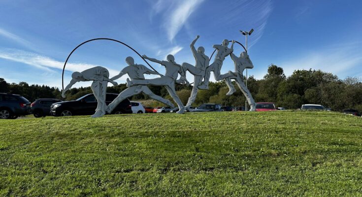 Statue of person bowling in multiple stages, and grass hill and in front of blue sky