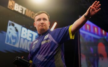 Darts player Luke Littler in a purple shirt hold up his left hand.