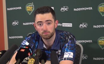 Luke Humphries in a Press conference, sitting staring into the frame. Green background with sponsorships, and several microphones placed in front of him