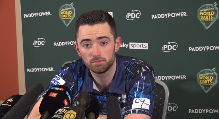 Luke Humphries in a Press conference, sitting staring into the frame. Green background with sponsorships, and several microphones placed in front of him