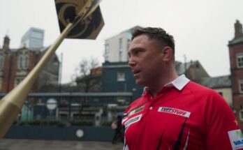 Gerwyn Price stands on the right hand side of the frame, with a golden dart statue to his right.