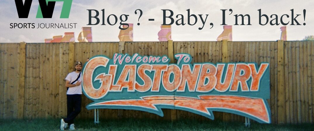 Hewlett-Me Write A Blog? - Baby, I'm back! Will Hewlett stood next to an orange and green 'Glastonbury' sign.