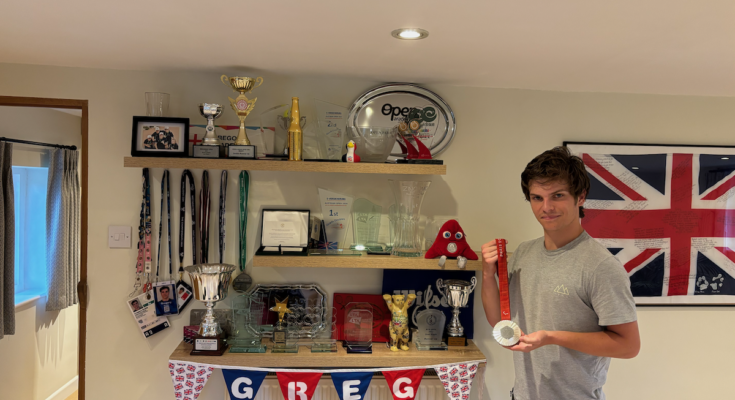 Wheelchair tennis player Greg Slade holding his Paralympic silver medal to the right, next to three shelves of accolades.