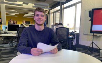 The presenter, Charlie Stevens, sat down at a table holding a piece of paper whilst smiling.