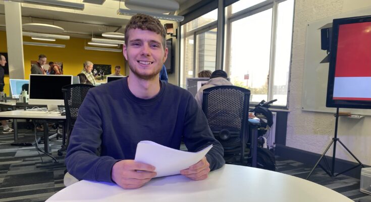 The presenter, Charlie Stevens, sat down at a table holding a piece of paper whilst smiling.