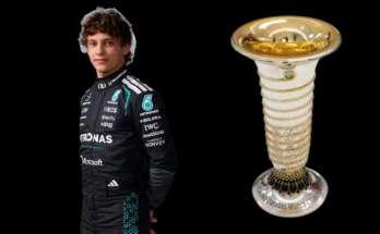 Driver in black racing suit posing with a trophy to the right of them.